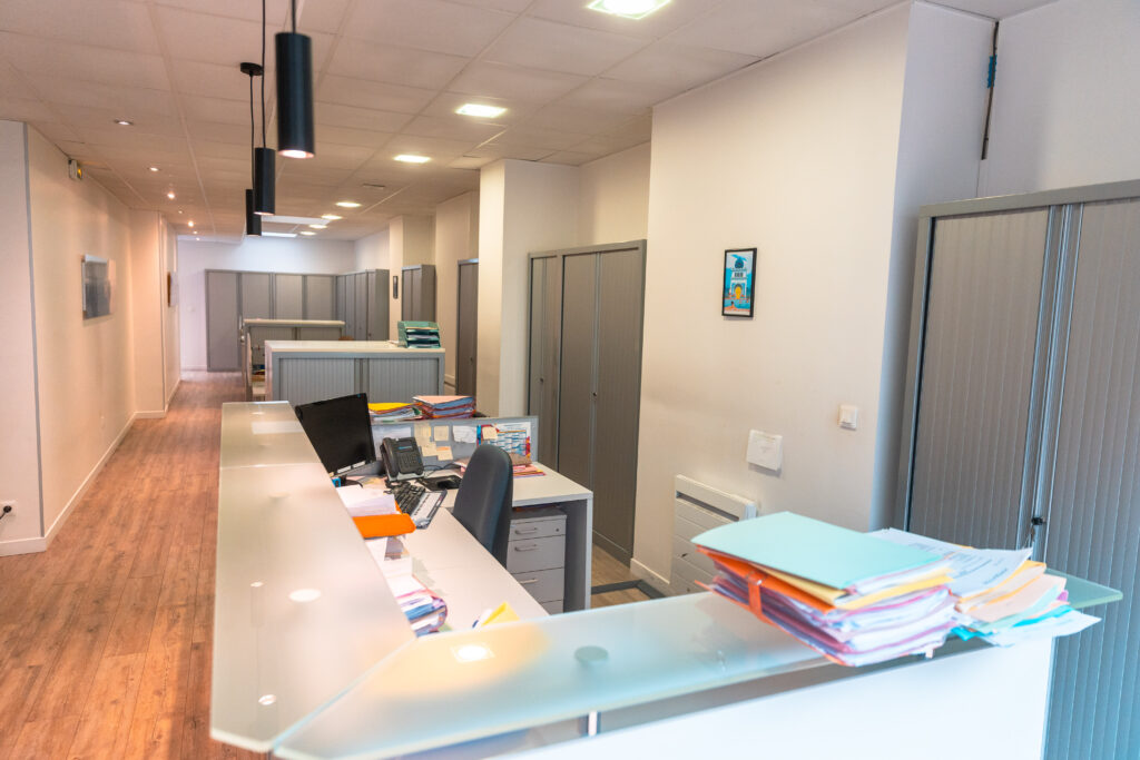 Un espace de bureau moderne et bien organisé. On y voit un couloir avec un sol en bois et des luminaires au plafond. Sur la droite, un bureau de réception avec un ordinateur, un téléphone et des dossiers empilés. À l'arrière-plan, des armoires de rangement et des postes de travail sont visibles, créant une ambiance professionnelle.